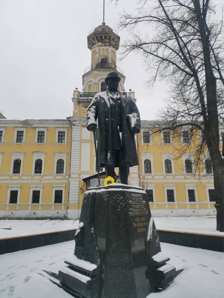 Памятник Аркадию Францевичу Кошко перед желто-белым зданием с башней в снегу