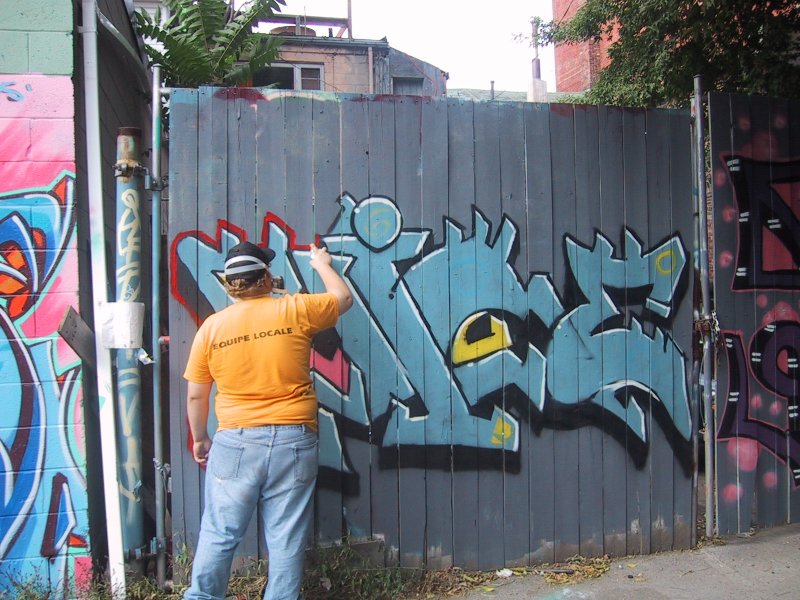 416 Graffiti Expo, Канада: художник в оранжевой футболке с надписью «EQUIPE LOCALE» рисует на деревянном заборе