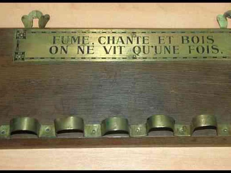 Деревянная табличка с бронзовыми крючьями и надписью «FUME, CHANTE ET BOIS ON NE VIT QU'UNE FOIS.»