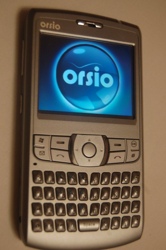 Orsio P745 на стене — серебристый смартфон с QWERTY-клавиатурой и логотипом в центре экрана