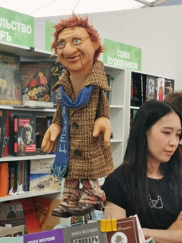 Bookfest 2025: кукла-малец в твидовом пальто у стеллажа с книгами