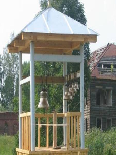 Деревянная колокольня с крестом и колоколами на фоне старого дома