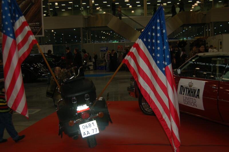 Мотоцикл на красной дорожке с двумя американским флагами и красным автомобилем