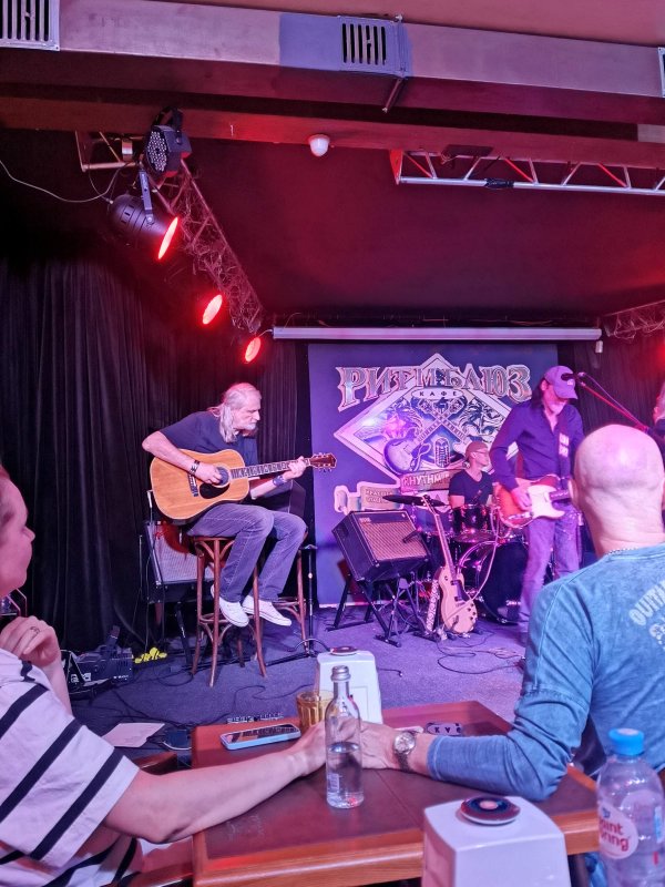 Ritm Blues Cafe в России: гитарист на стуле, барабанщик и бас-гитарист на сцене
