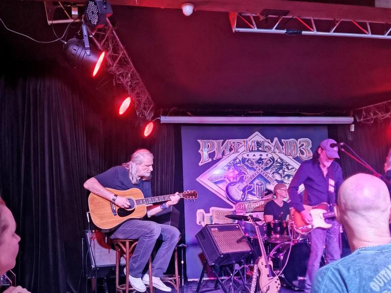 Ritm Blues Cafe в России: гитарист на стуле, барабанщик и бас-гитарист на сцене