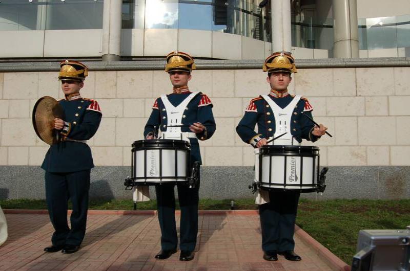 Три музыканта в парадной форме с барабанами и тарелкой на фоне современного здания