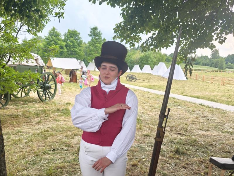 Times And Epochs 2026: женщина в красном жилете и цилиндре у военного стола